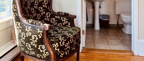 A room with a wooden floor, a patterned armchair, a doorway leading to a bathroom with a sink, toilet, and mirror.