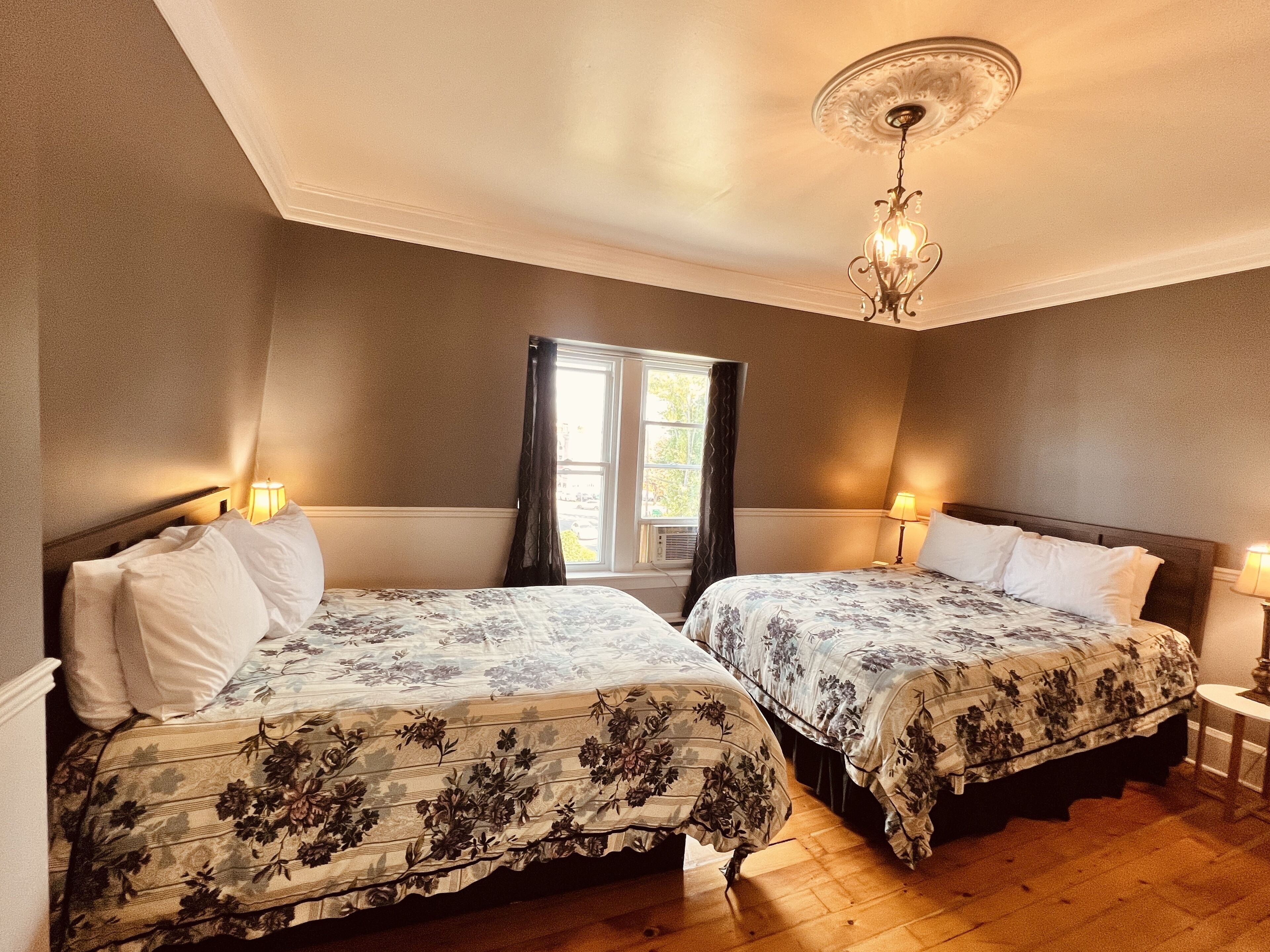 A hotel room with two beds, a window with curtains, a chandelier, and a small table with a lamp.