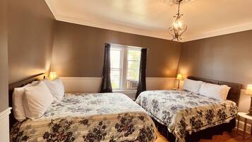 A hotel room with two beds, a window with curtains, a chandelier, and a small table with a lamp.