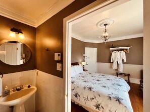Habitación de hotel con cama, mesita de noche, espejo, lámpara de pared, candelabro y baño con lavabo y espejo.