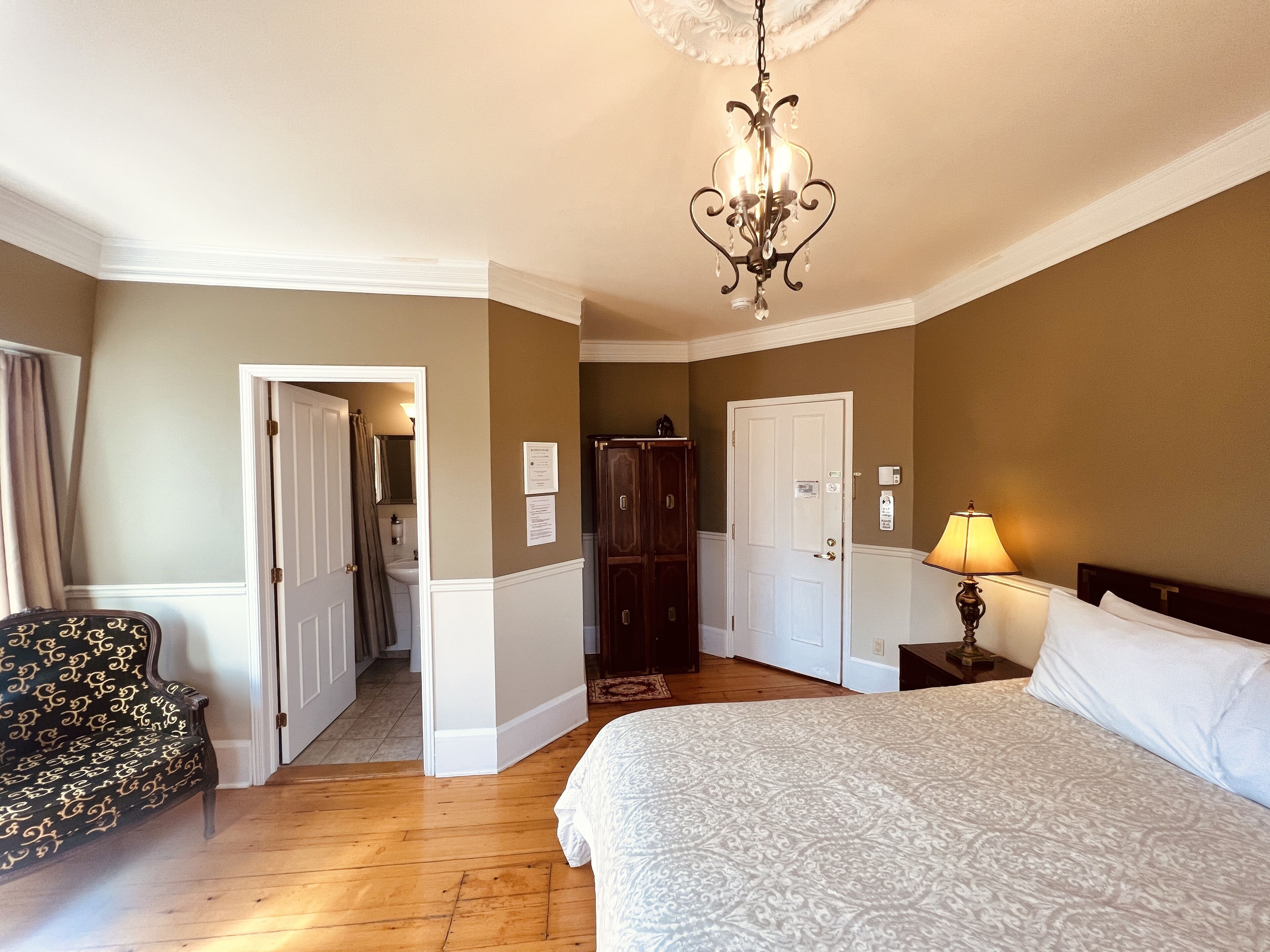 A hotel room with a bed, a chair, a desk with a lamp, a wardrobe, and a chandelier.
