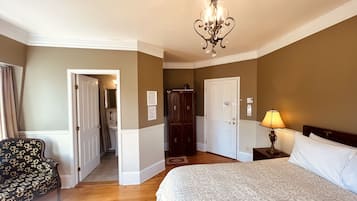 A hotel room with a bed, a chair, a desk with a lamp, a wardrobe, and a chandelier.