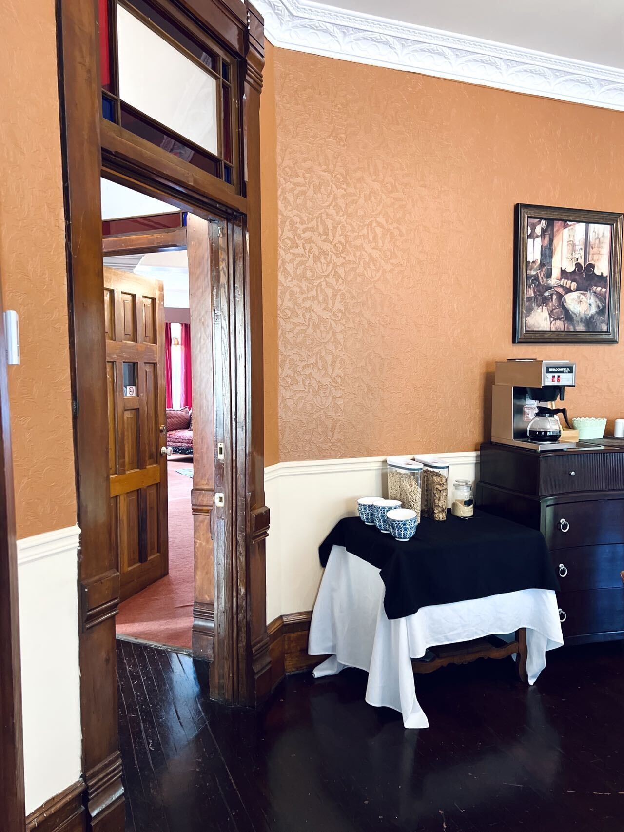 Une pièce avec une porte en bois, une table recouverte d’une nappe blanche et une machine à café posée sur une commode.