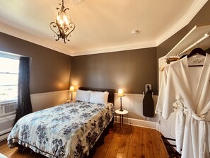 Un dormitorio con cama, dos lámparas de noche, un candelabro, un perchero y una ventana con cortinas.