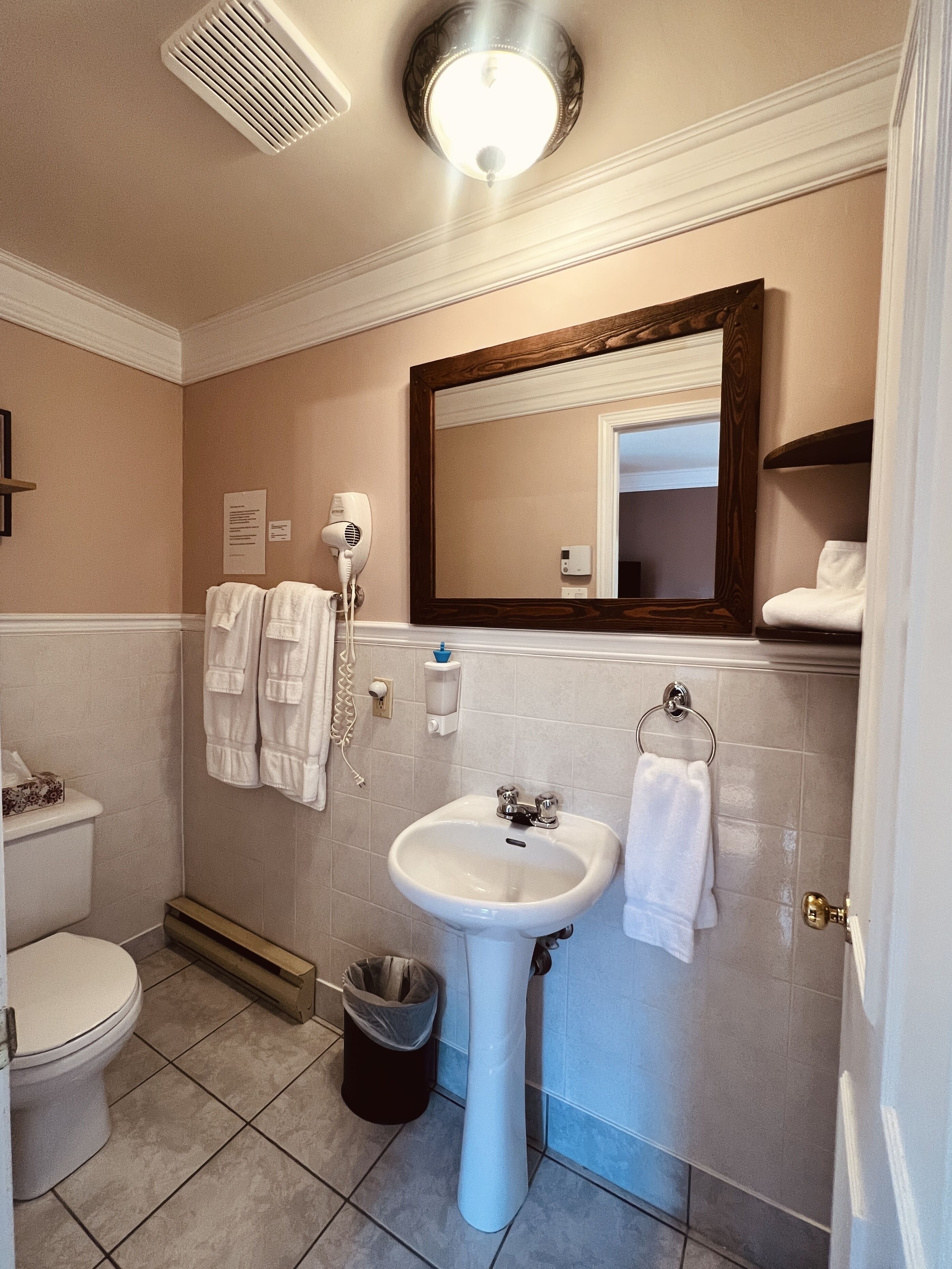 Ein Badezimmer mit Toilette, Waschbecken und Spiegel. Handtücher hängen an der Wand und ein Mülleimer steht bereit.