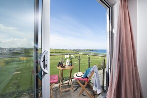 A balcony with a view of a golf course and the sea, a table with a bottle and glasses, and a chair with a hat and a blanket.
