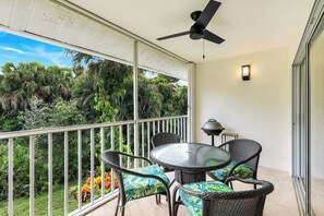 A balcony with a glass table and wicker chairs, a ceiling fan, and a view of lush greenery. - Sapphire Lakes Topaz (Naples)