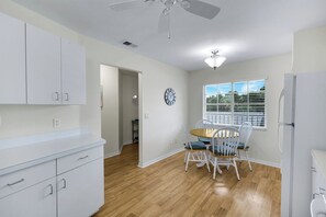 A kitchen with white cabinets, a dining table with chairs, a ceiling fan, and a window with blinds. - Sapphire Lakes Topaz (Naples)