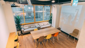 A modern office space with a long wooden table, colorful chairs, a sofa, and a large window with a city view. - Tokyo Guesthouse HIVE (Tokyo)