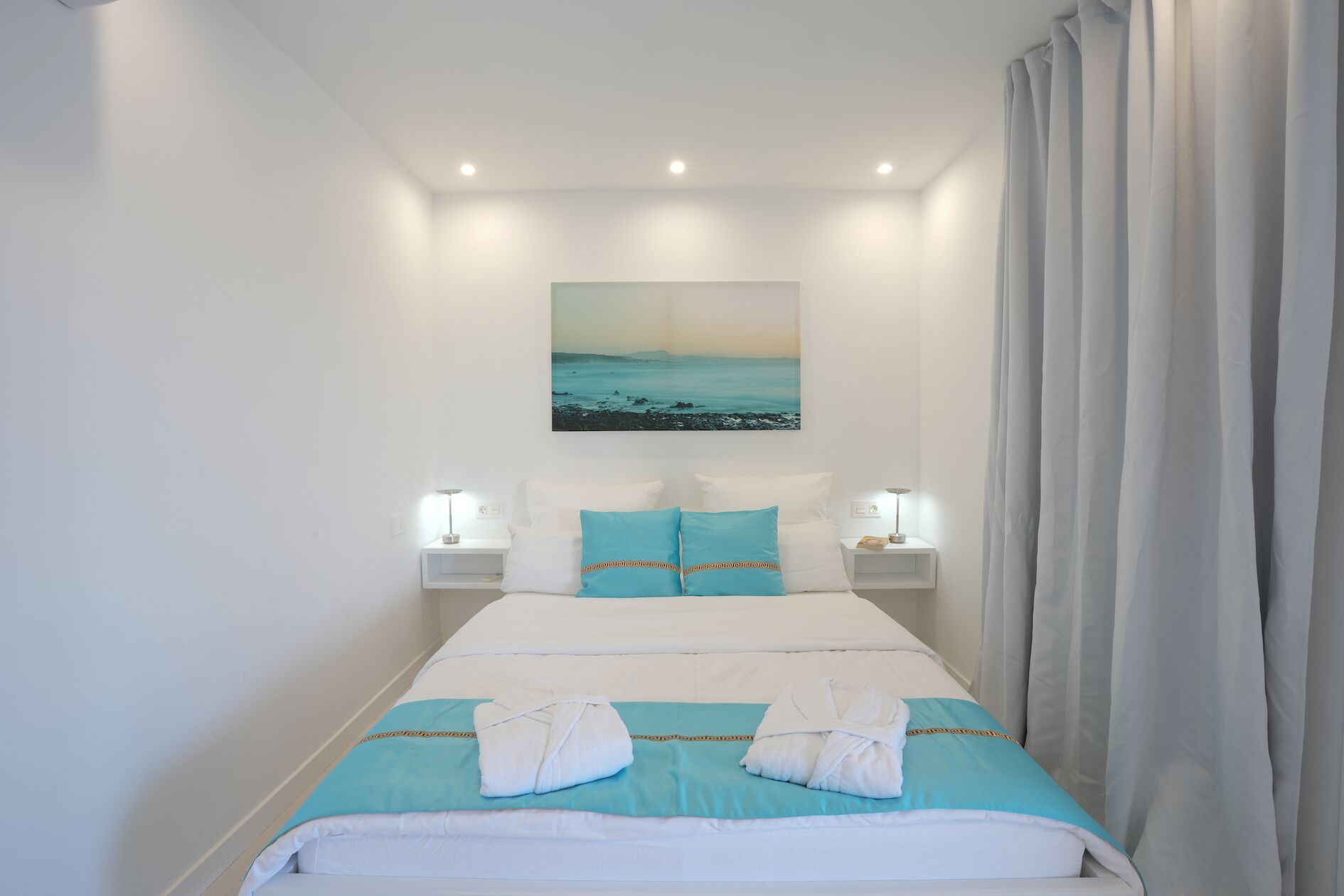 Une chambre d’hôtel avec un lit, deux tables de chevet, un tableau représentant une scène de plage et un rideau.