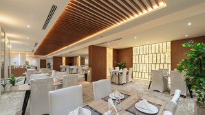 A spacious hotel dining area with multiple tables set for dining, wooden paneling on the ceiling, and potted plants. - Ross Hotel  (Riyadh)