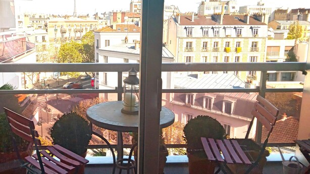 A balcony with a round table and two chairs, overlooking a cityscape with various buildings and a prominent tower.