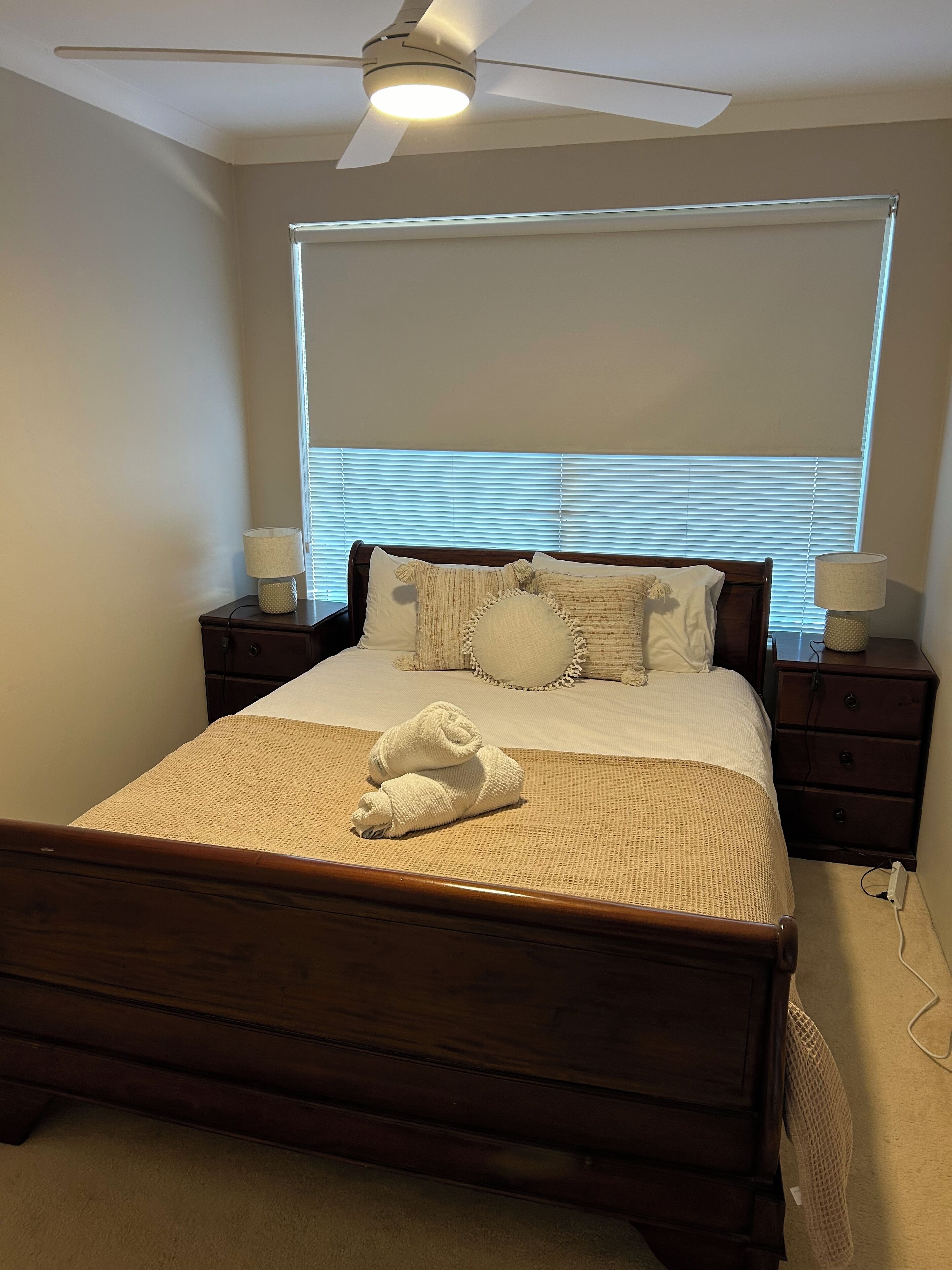 Une chambre à coucher comprenant un lit, deux tables de chevet, un ventilateur de plafond et une fenêtre avec des stores.