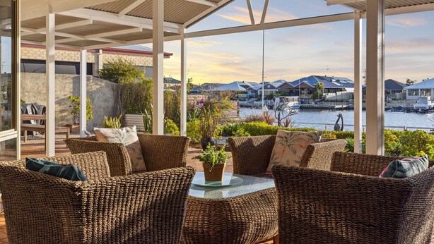 A covered outdoor seating area with wicker furniture, a glass table, and a view of a marina with boats and houses in the background.