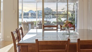 A dining area with a glass table and wooden chairs, a vase with flowers, and a view of a body of water and buildings through large windows.