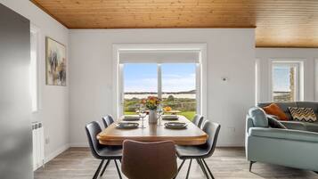 A dining area with a wooden table and chairs, a view of the outdoors, and a sofa in the background.