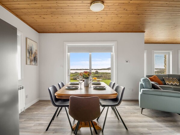 A dining area with a wooden table and chairs, a view of the outdoors, and a modern living space with a sofa and cushions.