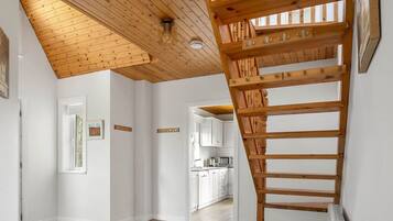 Habitación amplia con techo de madera, una escalera y una cocina visible a través de una puerta abierta.