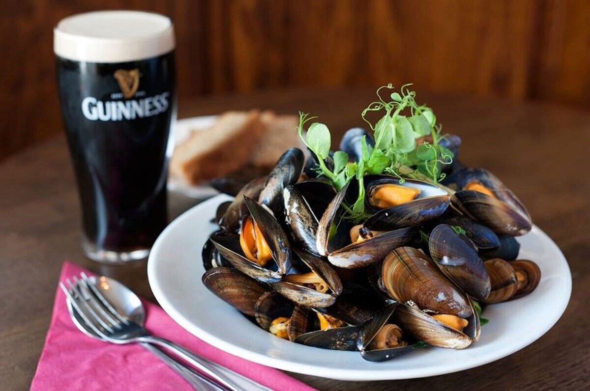 A plate of mussels with a glass of Guinness beer and a fork on a table.