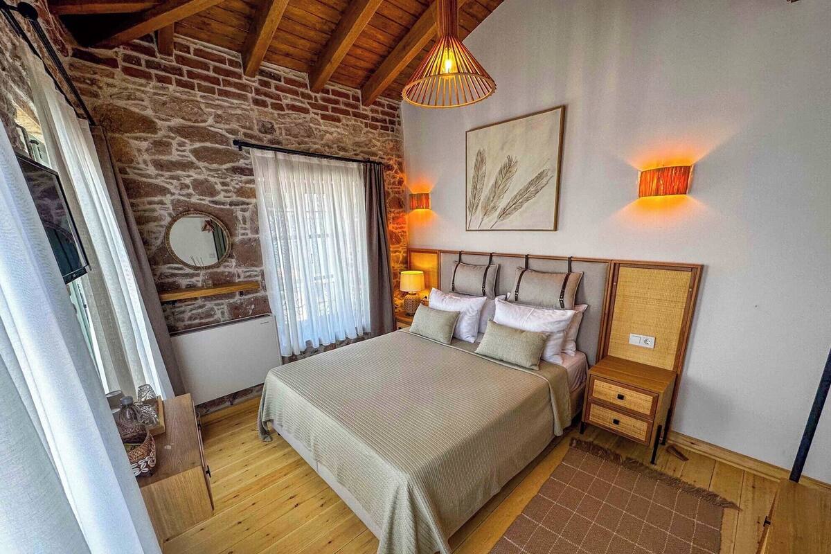 A bedroom with a stone wall, wooden ceiling beams, a bed with pillows, bedside lamps, and a framed artwork.