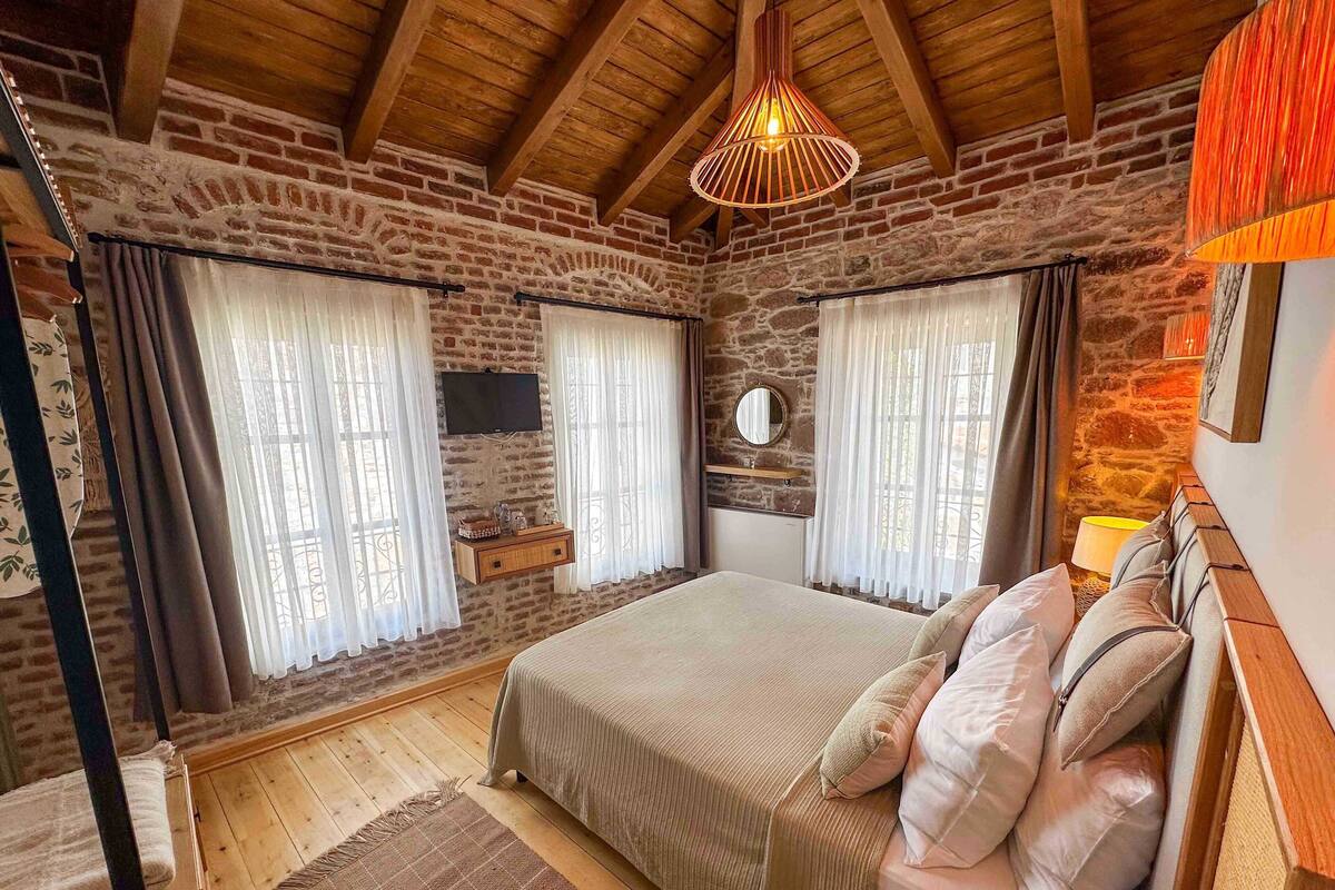 A bedroom with a stone wall, wooden ceiling beams, a bed with a headboard, a bedside table, and a hanging light fixture.