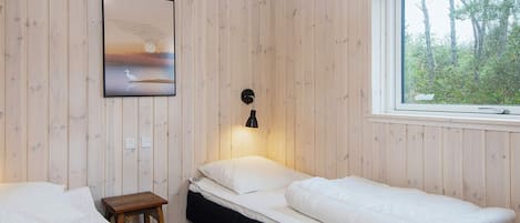 A room with two beds, a small table, a wall-mounted lamp, and a window with a view of trees.