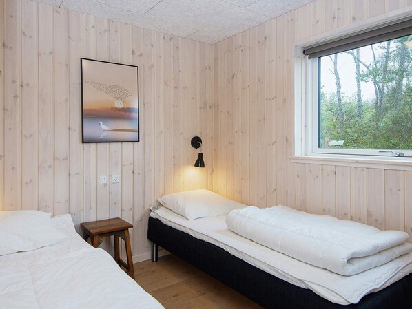 A room with two beds, a small table, a wall-mounted lamp, and a window with a view of trees.
