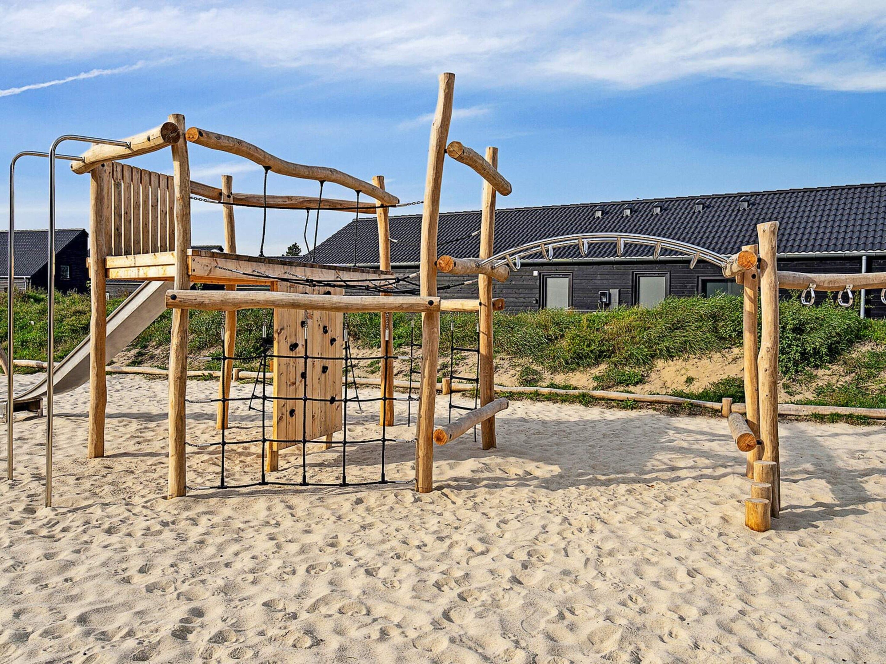 Una estructura de juego de madera en una playa arenosa, con un edificio al fondo.