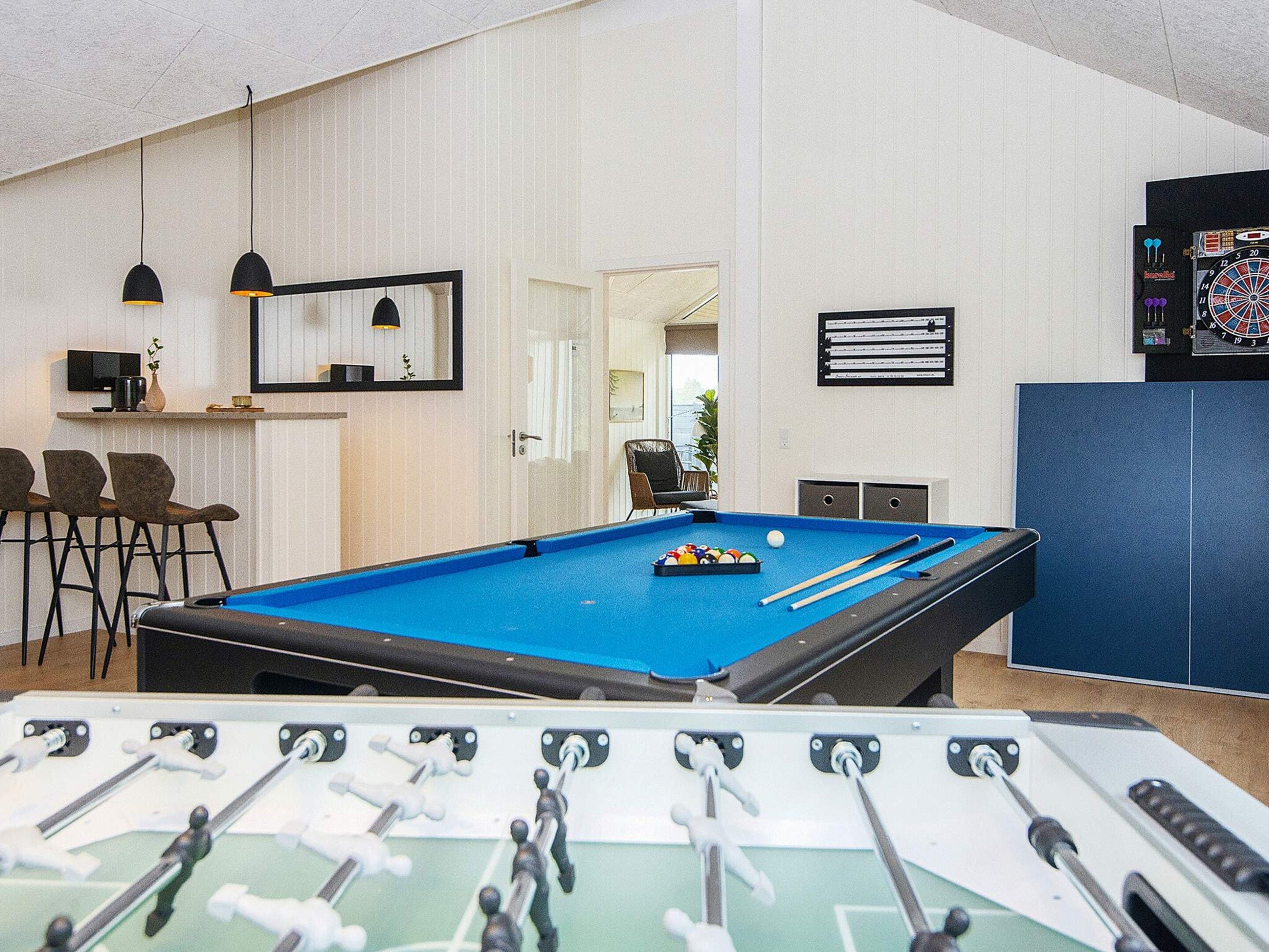 A pool table with a foosball table in the foreground, a dartboard, and a bar area with seating.