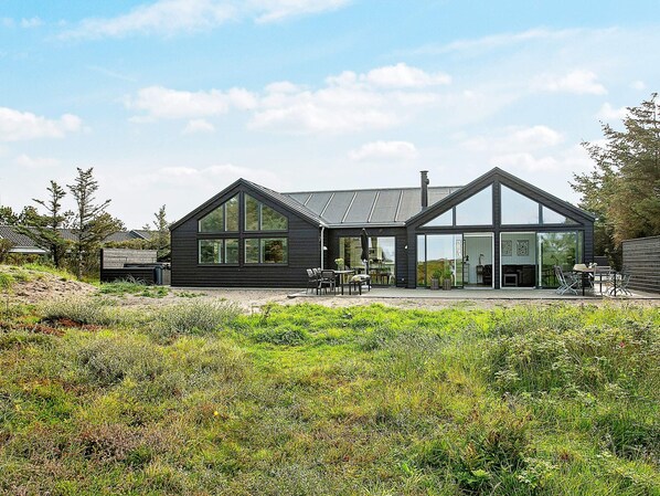 A modern two-story house with a large glass facade, a sloped roof, and a patio area with outdoor furniture.