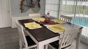 Una zona de menjador amb una taula de fusta fosca, cadires blanques i roba de taula groga. Hi ha un objecte decoratiu penjant a la paret i una ampolla sobre la taula.