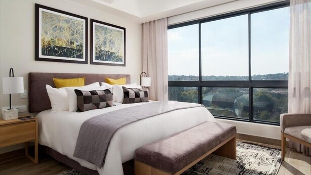 A modern bedroom with a large bed, bedside tables, a bench, and a view of the city through large windows.