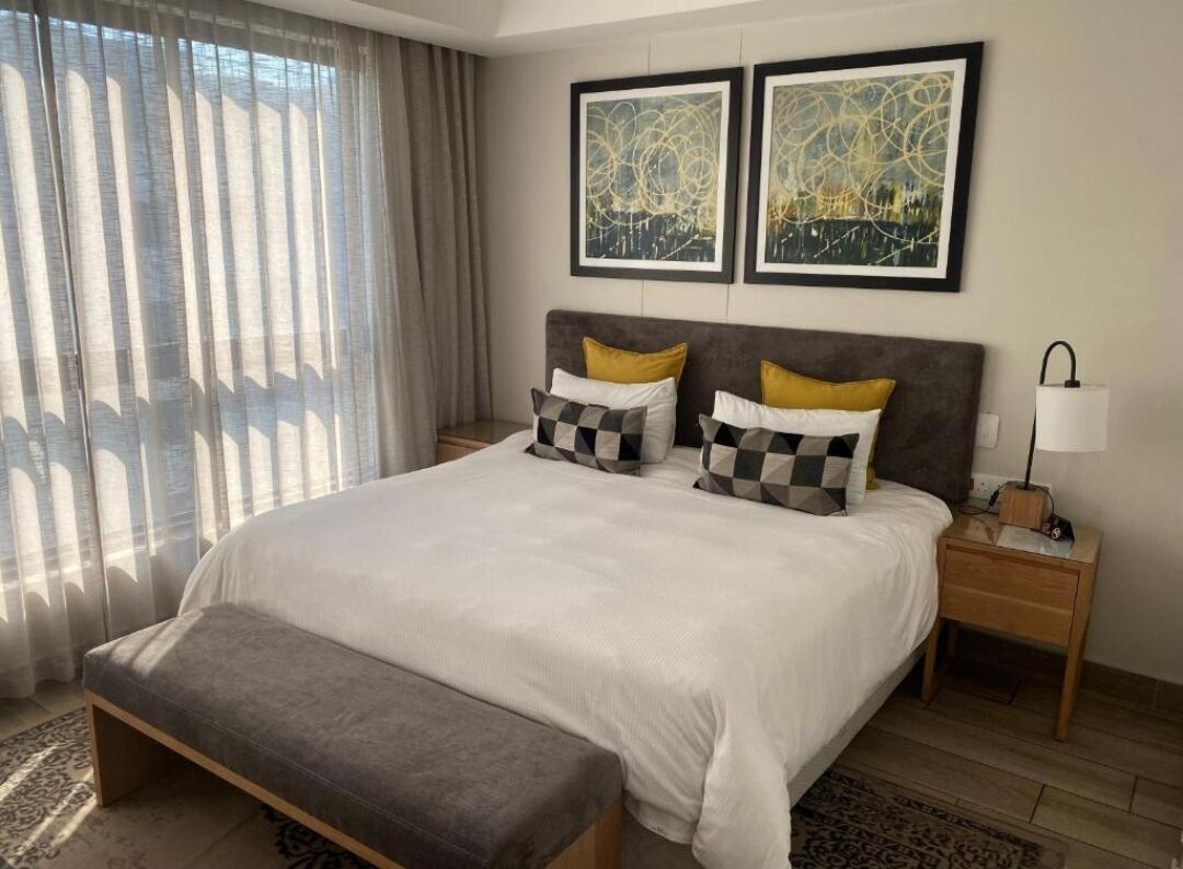 Un dormitorio ordenado con una cama grande, dos mesitas de noche, un banco y una ventana con cortinas.
