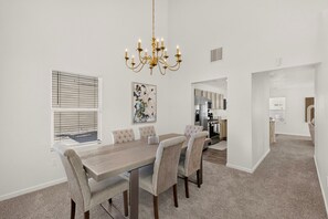 A dining area with a wooden table and upholstered chairs, a chandelier, a window with blinds, and a kitchen in the background. - Peaceful 3bd/2.5ba home w/ yard, garage (Albuquerque)