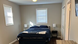A bedroom with a bed, two bedside tables, a wooden dresser, and two windows with blinds.