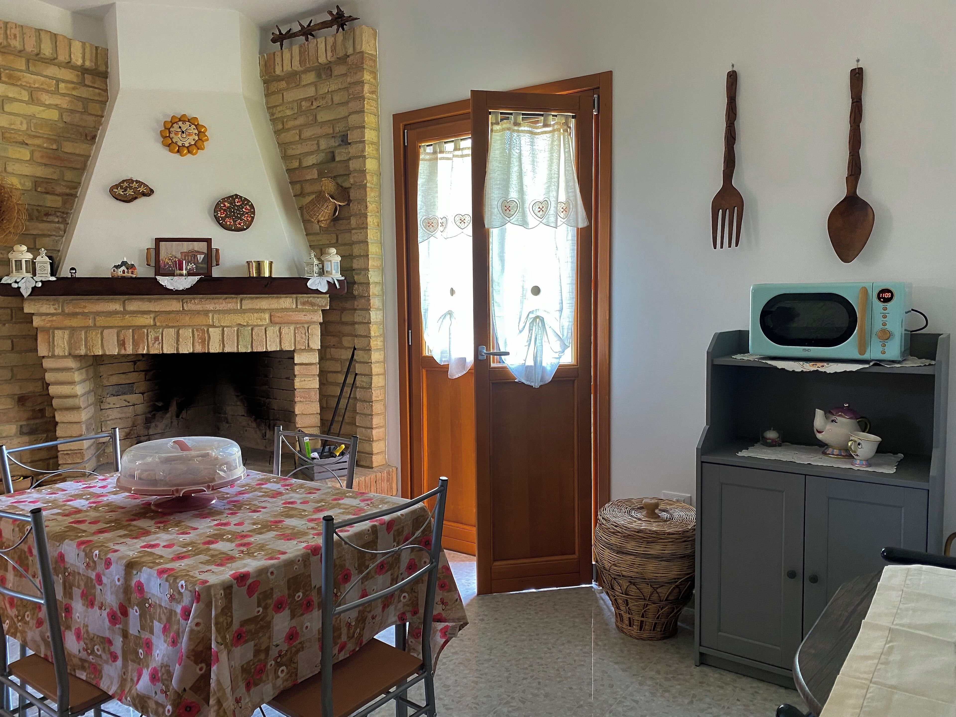 A cozy room with a brick fireplace, a dining table with a floral tablecloth, a wicker basket, and a microwave on a shelf.