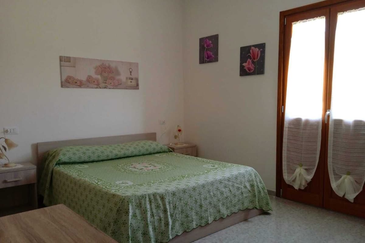 A bedroom with a bed, a nightstand, a wooden table, and a door with curtains.