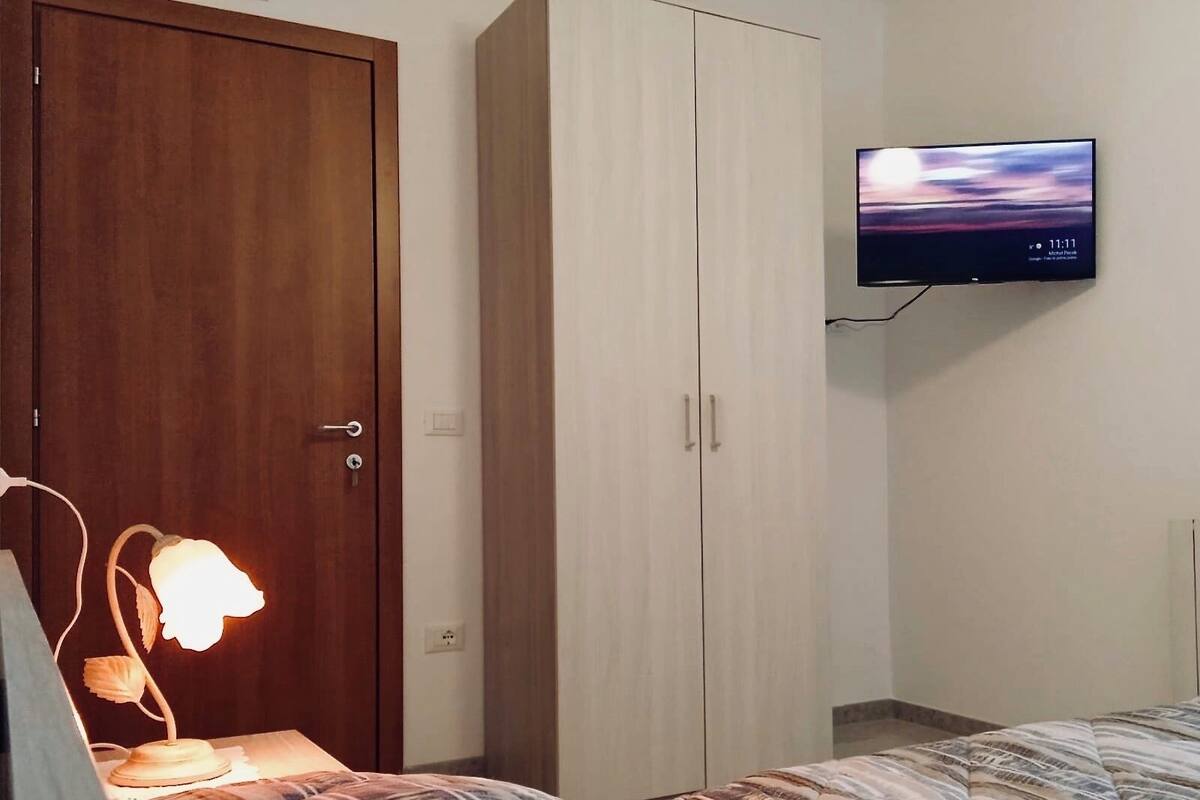 A bedroom with a bed, a nightstand, a lamp, a wardrobe, and a TV mounted on the wall.