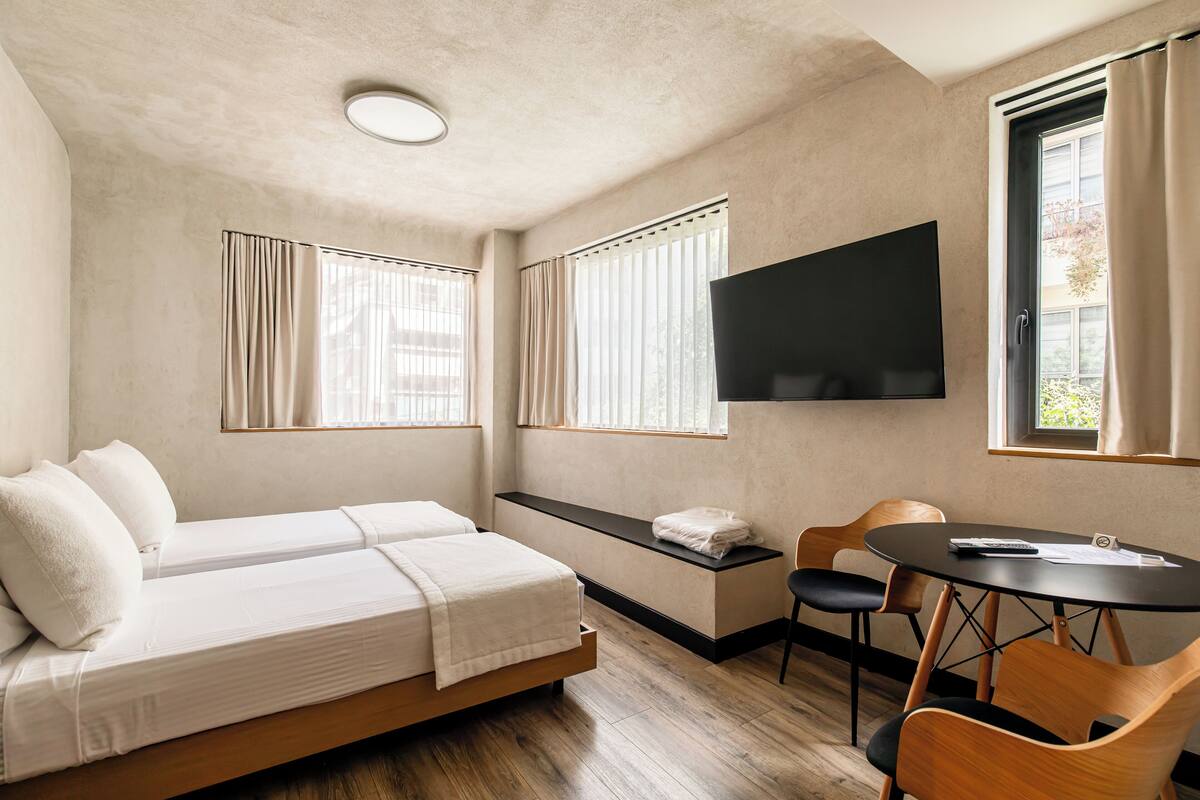Une chambre comprenant un lit, un bureau, une chaise, un téléviseur et deux fenêtres avec des rideaux.