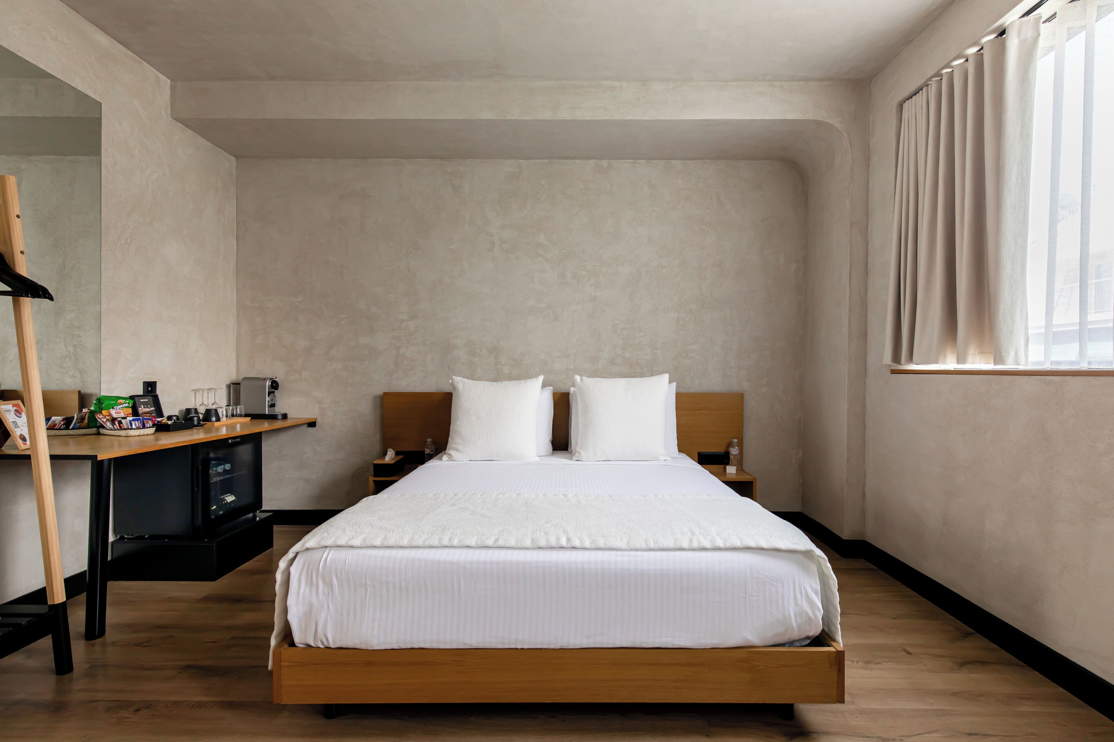 Una cama individual con estructura de madera, una mesita de noche con lámpara, un escritorio pequeño con silla, y una ventana con cortinas.