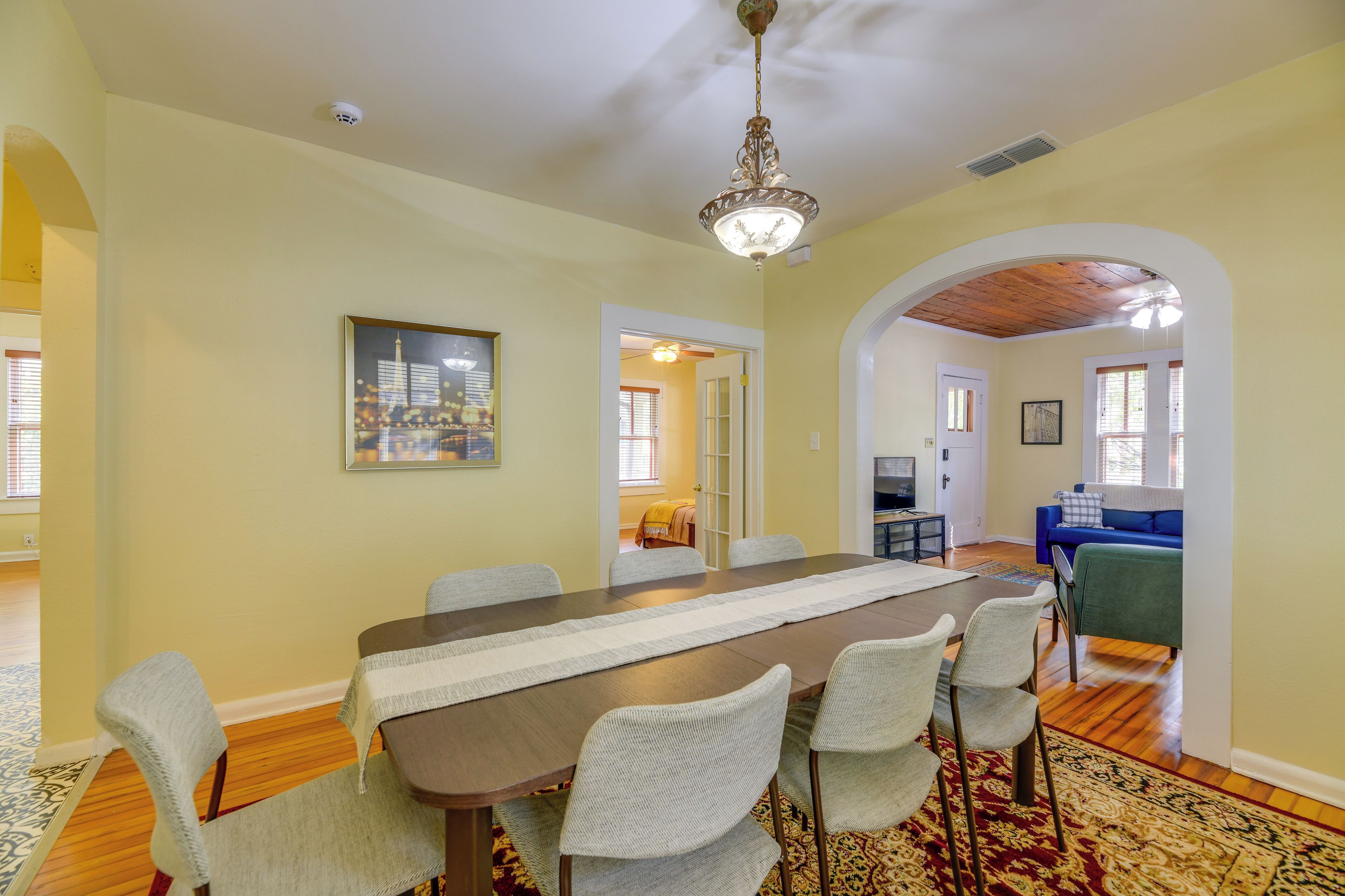 A dining area with a long table, chairs, and a chandelier. There is a doorway leading to another room with a blue couch and a television.