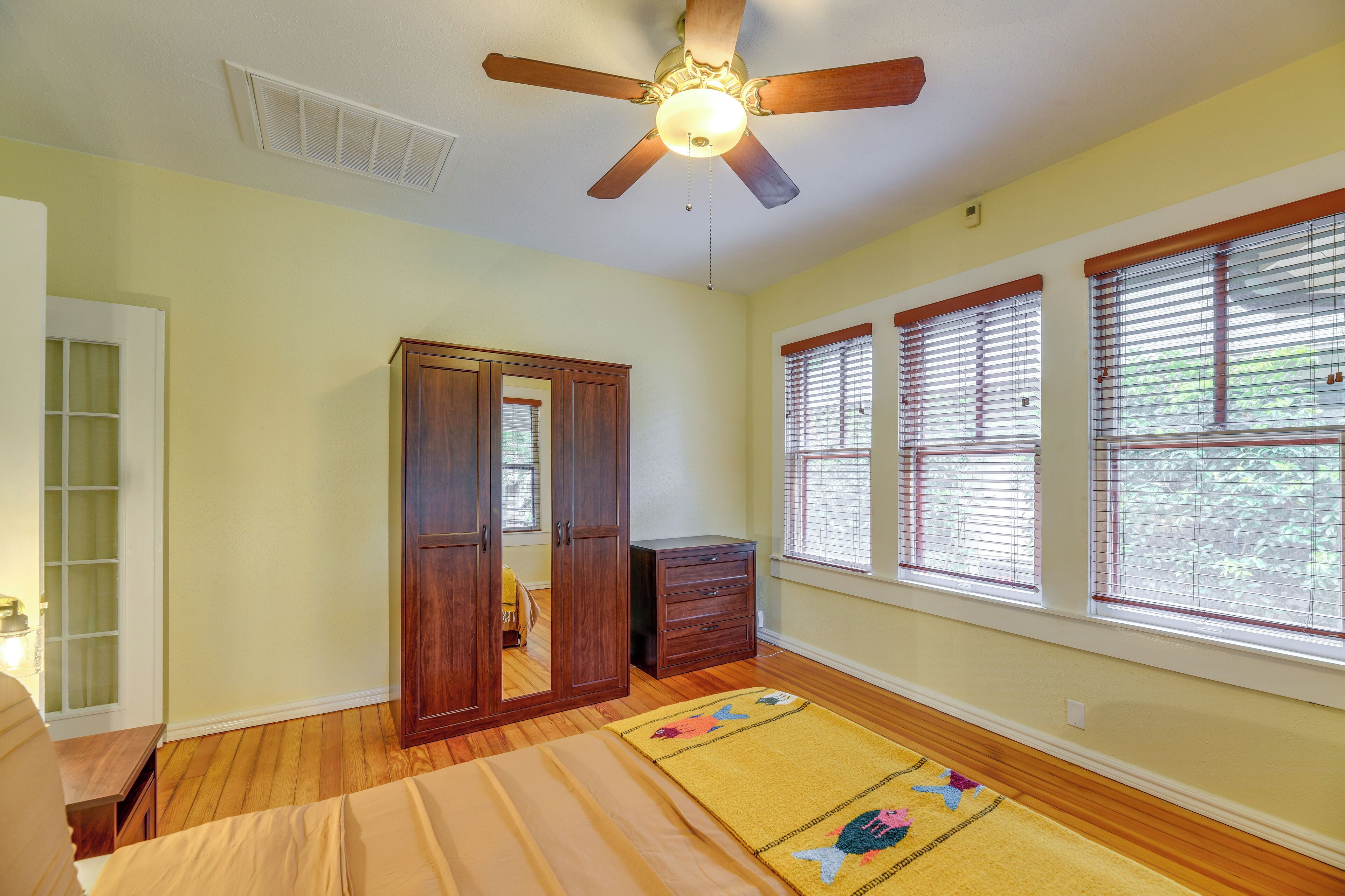 A bedroom with a bed, a ceiling fan, wooden wardrobe, and windows with blinds.