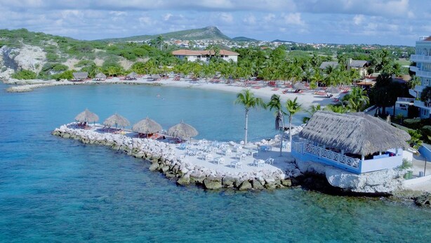 A coastal resort with a rocky pier, thatched-roof huts, and a clear blue sea. - Blue Bay Beach Apartments (Sint Michiel)