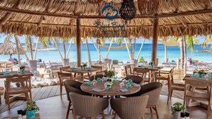 A beachside restaurant with thatched roofs, wicker furniture, and tables set for dining. - Blue Bay Beach Apartments (Sint Michiel)