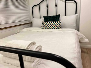 A neatly made bed with white linens and a checkered pillow, a black metal headboard, and a window with blinds. - Essex 2-bed House nr Stanstead With Arcade Machine (Braintree)