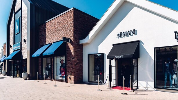A row of storefronts including a brick building and a white building with an 'ARMANI' sign. - Essex 2-bed House nr Stanstead With Arcade Machine (Braintree)