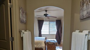 A hallway leading to a bedroom with a bed, a chair, a ceiling fan, and a window with curtains.