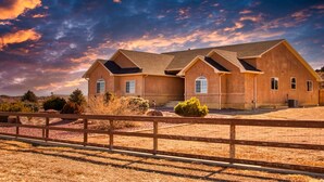 A large, single-story house with a sloped roof and multiple windows, surrounded by a wooden fence and dry grass.