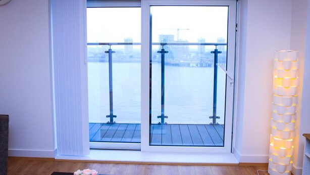 A room with a large sliding glass door leading to a balcony with a view of a body of water and buildings. There is a clock on the wall and a tall, cylindrical light fixture on the right. - Thout Seaview Luxury Apartment (London)