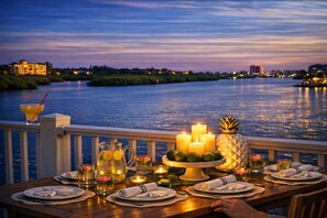 Ein Esstisch, gedeckt für ein Abendessen, mit Kerzen, einer Ananas und einem Getränk in einer Karaffe, mit Blick auf einen Fluss im Abenddämmerung.